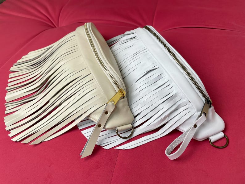 May include: Two fringed leather fanny packs in cream and white, with zip closures and strap details. The cream-coloured bag has a gold zip, while the white bag has a silver zip. Both bags have a D-ring attachment.