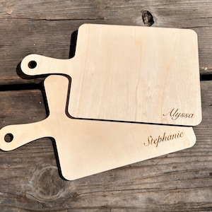 May include: Two wooden cutting boards with handles. The top board has the name "Alyssa" engraved on it. The bottom board has the name "Stephanie" engraved on it.