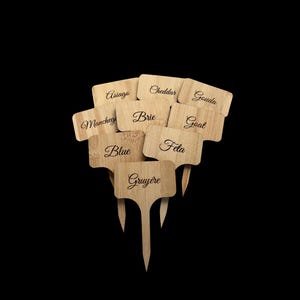 May include: A collection of wooden cheese markers with pointed stakes. Each marker is engraved with a different cheese name in elegant script, including Asiago, Cheddar, Gouda, Manchego, Brie, Goat, Blue, Feta, and Gruyère. The markers are light brown against a black background.
