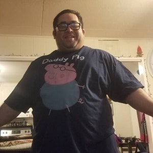 May include: A man wearing a navy blue t-shirt with a pink cartoon pig wearing glasses and the text "Daddy Pig".