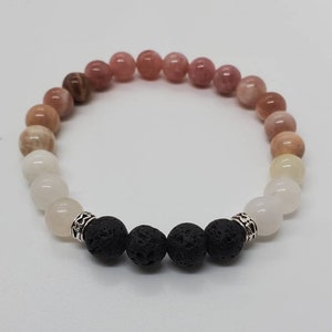 May include: A bracelet made of beads in shades of white, peach, and brown. The bracelet includes a section of black lava beads, separated by silver-coloured spacers. The bracelet is displayed against a white background. The beads are approximately 0.8 cm in diameter.