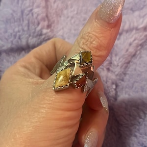 May include: A silver ring with multiple geometric shapes and inlaid gemstones. The ring features a square, a diamond, and a rectangular stone in shades of brown, yellow, and red. Small silver accents and a star-shaped element add detail.
