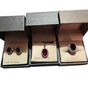 May include: Three jewelry boxes, each containing a piece of jewelry. The boxes hold a pair of earrings, a pendant necklace, and a ring. Each piece features a large, oval, red stone surrounded by smaller, clear stones. The text "Cubic Zirconia" is visible.
