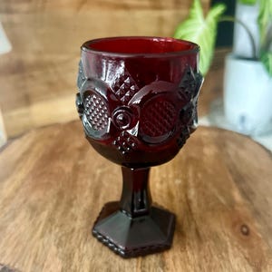 Puede incluir: Una copa de cristal rojo rubí vintage con detalles ornamentados. La copa presenta un patrón decorativo alrededor del cuenco y una base hexagonal. El cristal es de un color rojo intenso y rico, y la copa está sobre una superficie de madera.