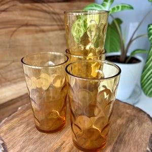 Puede incluir: Cuatro vasos de color ámbar con un patrón texturizado en forma de burbujas. Los vasos están colocados sobre una tabla de servir de madera, mostrando sus tonos cálidos y acogedores. La cristalería es ideal para servir bebidas.