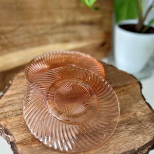 Puede incluir: Tres platos de vidrio rosa con un diseño acanalado están apilados sobre una tabla de servir de madera. Los platos tienen forma circular y un centro ligeramente elevado. La tabla de madera tiene un borde natural y orgánico.