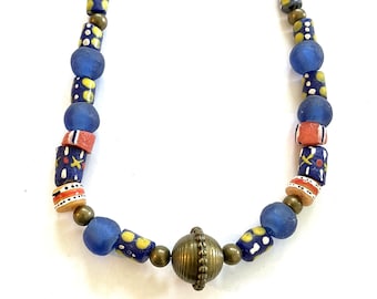May include: A necklace featuring a central round brass bead with a ribbed texture, flanked by smaller brass spheres. The strand includes matte blue glass beads, cylindrical beads with yellow and white patterns, and orange beads with black and white stripes.