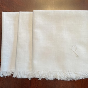 May include: Three white linen napkins with fringed edges. The napkin on the right has an embroidered letter 'P' in the corner.