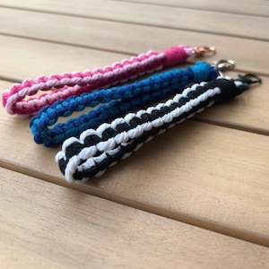 May include: Three handmade wrist lanyards in pink, blue, and black and white. Each lanyard is made of braided cord and has a metal clasp. The lanyards are lying on a light brown wooden surface.