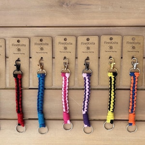 May include: A selection of macrame keyrings in a variety of colours, including green, black and white, red, blue, pink, purple, yellow, orange, and red and white. Each keyring has a metal clasp and a keyring, and is attached to a cardboard tag.