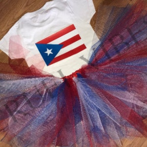 May include: A white t-shirt with a red, white, and blue Puerto Rican flag design on the back. The shirt is paired with a red, white, and blue tulle tutu.