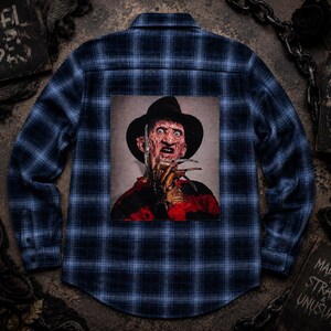 May include: A blue and black plaid long-sleeve button-down shirt featuring a large square graphic on the back. The graphic depicts a character wearing a dark fedora hat and a red and black striped sweater, with a metal clawed glove held near the face.