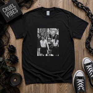 May include: A black short-sleeved t-shirt featuring a central black and white photographic print of four individuals standing together. The garment has a crew neckline and a standard fit. The graphic depicts people in light shirts and dark skirts.