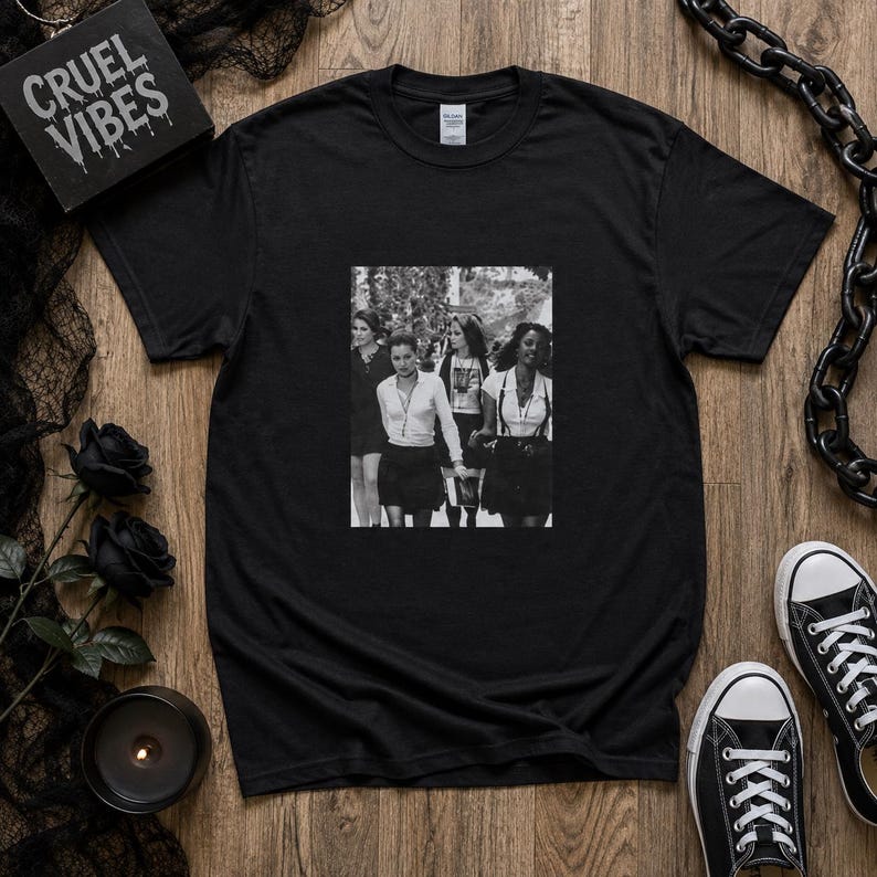 May include: A black short-sleeved t-shirt featuring a central black and white photographic print of four individuals standing together. The garment has a crew neckline and a standard fit. The graphic depicts people in light shirts and dark skirts.