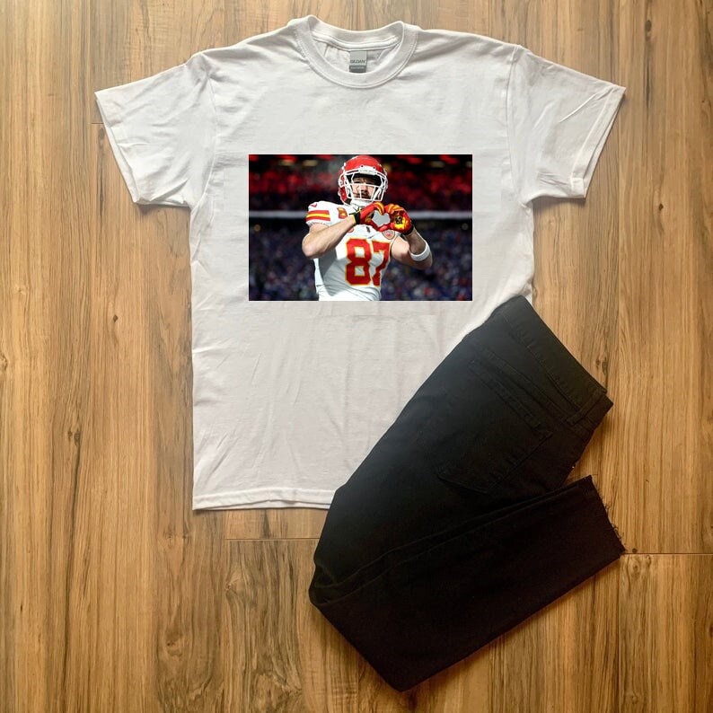 K&ouml;nnte beinhalten: Wei&szlig;es T-Shirt mit einem aufgedruckten Bild eines Footballspielers, der ein rot-wei&szlig;-gelbes Trikot mit der Nummer 87 tr&auml;gt. Der Spieler tr&auml;gt einen Helm und h&auml;lt einen Football.