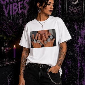 以下が含まれることがあります： 胸元に長方形のグラフィックプリントが施された白い半袖Tシャツ。プリントには、両手を上げた人物が指に複数の大きなシルバーリングをはめている様子が描かれています。Tシャツはクルーネックのデザインです。