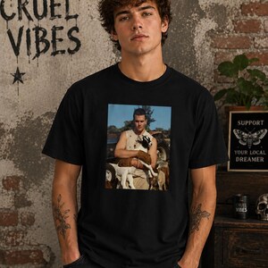 May include: A black short-sleeve t-shirt featuring a rectangular graphic print on the chest. The print depicts a person wearing a sleeveless top while holding a small black and white goat in an outdoor setting with a vehicle in the background.