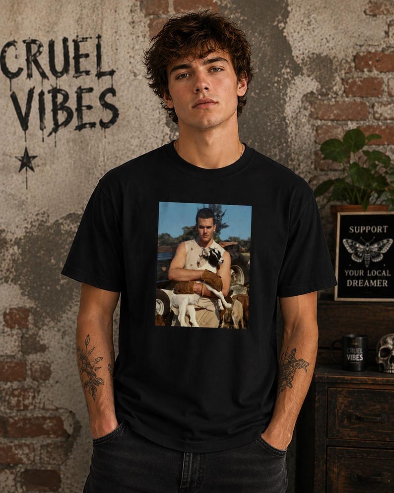 May include: A black short-sleeve t-shirt featuring a rectangular graphic print on the chest. The print depicts a person wearing a sleeveless top while holding a small black and white goat in an outdoor setting with a vehicle in the background.