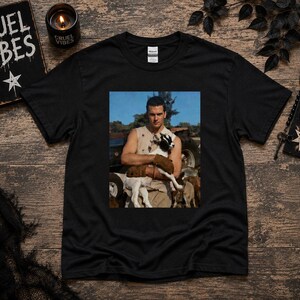 May include: A black short-sleeve t-shirt featuring a rectangular graphic print on the chest. The print shows a person wearing a sleeveless top and brown gloves, holding a small black and white goat in an outdoor setting with a tractor in the background.