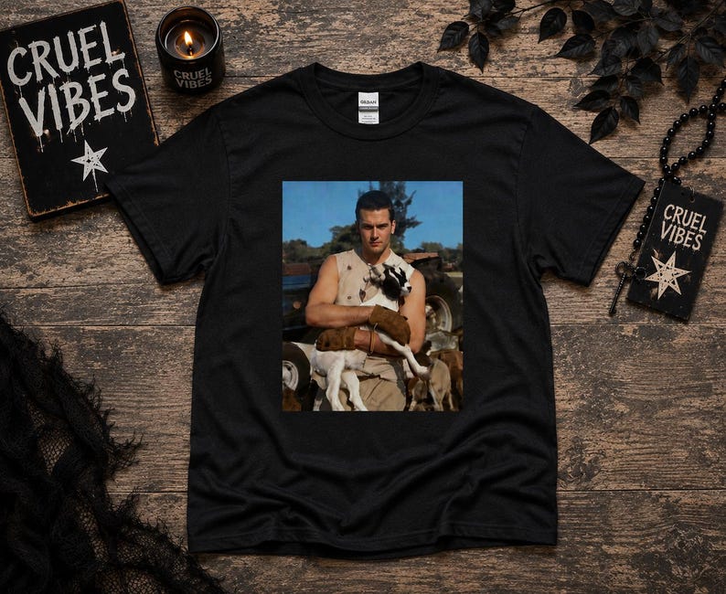 May include: A black short-sleeve t-shirt featuring a rectangular graphic print on the chest. The print shows a person wearing a sleeveless top and brown gloves, holding a small black and white goat in an outdoor setting with a tractor in the background.