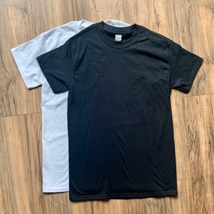 K&ouml;nnte beinhalten: Zwei schlichte T-Shirts, eines wei&szlig; und eines schwarz, flach auf einer Holzoberfl&auml;che gelegt.