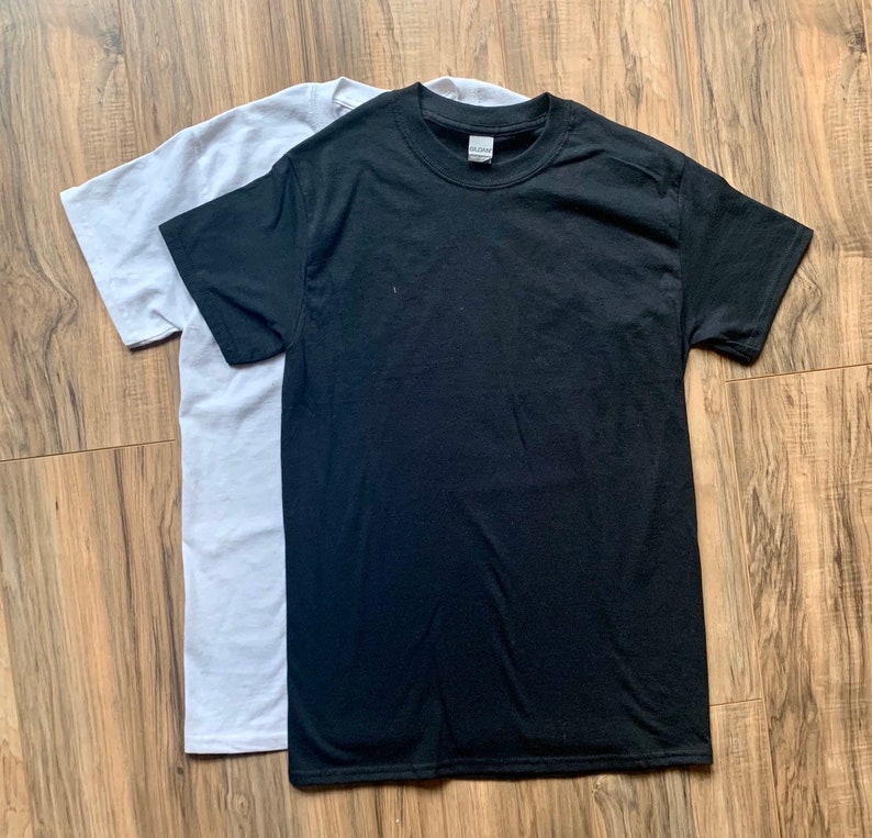 K&ouml;nnte beinhalten: Zwei schlichte T-Shirts, eines wei&szlig; und eines schwarz, flach auf einer Holzoberfl&auml;che gelegt.