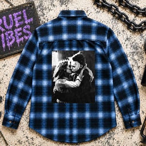 Puede incluir: Una camisa de manga larga con botones y un patr&oacute;n de cuadros en azul y negro. La parte posterior de la camisa muestra un estampado gr&aacute;fico en blanco y negro de una pareja abraz&aacute;ndose. La camisa tiene un cuello est&aacute;ndar y pu&ntilde;os abotonados.