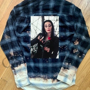 May include: A blue and black plaid flannel shirt with a faded bleach wash effect. A rectangular image of a woman in black holding a red rose is centered on the back of the shirt. The shirt has a button-down front and long sleeves.