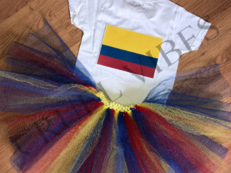 May include: A white t-shirt with a yellow, blue, and red Colombian flag design. The shirt is paired with a multi-colored tulle tutu skirt with yellow, red, blue, and gold layers.