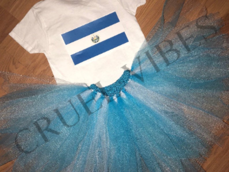 May include: A white t-shirt with the flag of El Salvador printed on it, paired with a blue and white tulle tutu skirt. The skirt has a sparkly finish and the text "CRUEL VIBES" is visible on the skirt.