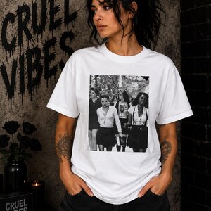 May include: A white short-sleeved t-shirt featuring a black and white rectangular graphic print on the chest. The graphic depicts four individuals standing together in a group. The shirt has a crew neckline and a standard fit, constructed from cotton fabric.