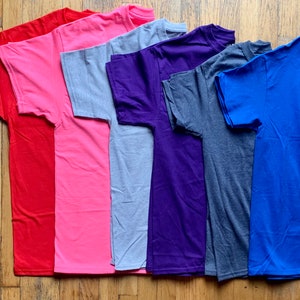 K&ouml;nnte beinhalten: Sieben einfarbige T-Shirts in verschiedenen Farben, darunter Rot, Rosa, Grau, Lila, Dunkelgrau und Blau. Die T-Shirts sind gefaltet und auf einer Holzoberfl&auml;che ausgelegt.