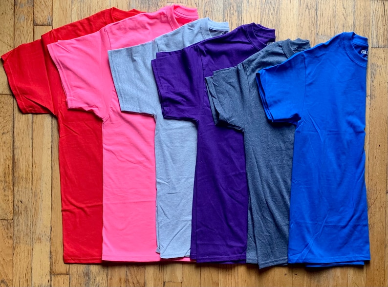 K&ouml;nnte beinhalten: Sieben einfarbige T-Shirts in verschiedenen Farben, darunter Rot, Rosa, Grau, Lila, Dunkelgrau und Blau. Die T-Shirts sind gefaltet und auf einer Holzoberfl&auml;che ausgelegt.