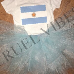 May include: A white baby onesie with a blue and white Argentinian flag design. The onesie is paired with a light blue tulle tutu.