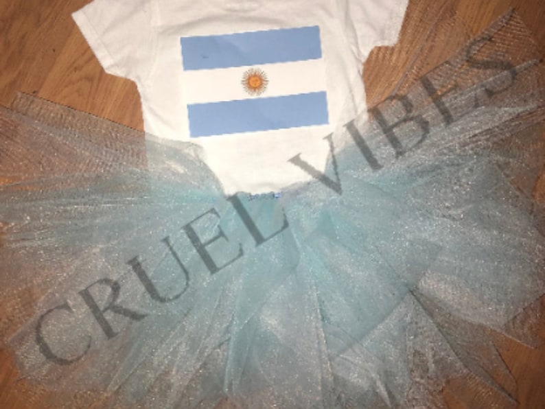 May include: A white baby onesie with a blue and white Argentinian flag design. The onesie is paired with a light blue tulle tutu.