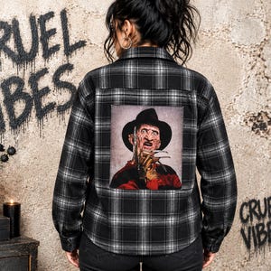 May include: A black and white plaid button-down shirt featuring a large rectangular graphic on the back. The graphic depicts a character wearing a brown fedora and a red and black striped sweater, with metallic clawed fingers raised near the face.