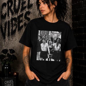 May include: A black short-sleeved t-shirt featuring a rectangular black and white photographic print on the chest. The graphic depicts four individuals in school-style attire, including button-down shirts, skirts, and suspenders, standing together.