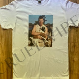 Puede incluir: Camiseta blanca con una imagen de estilo vintage de una persona sosteniendo una cabra. La imagen est&aacute; centrada en la camiseta y tiene una apariencia descolorida y envejecida. La camiseta est&aacute; hecha de una tela suave y de color claro.