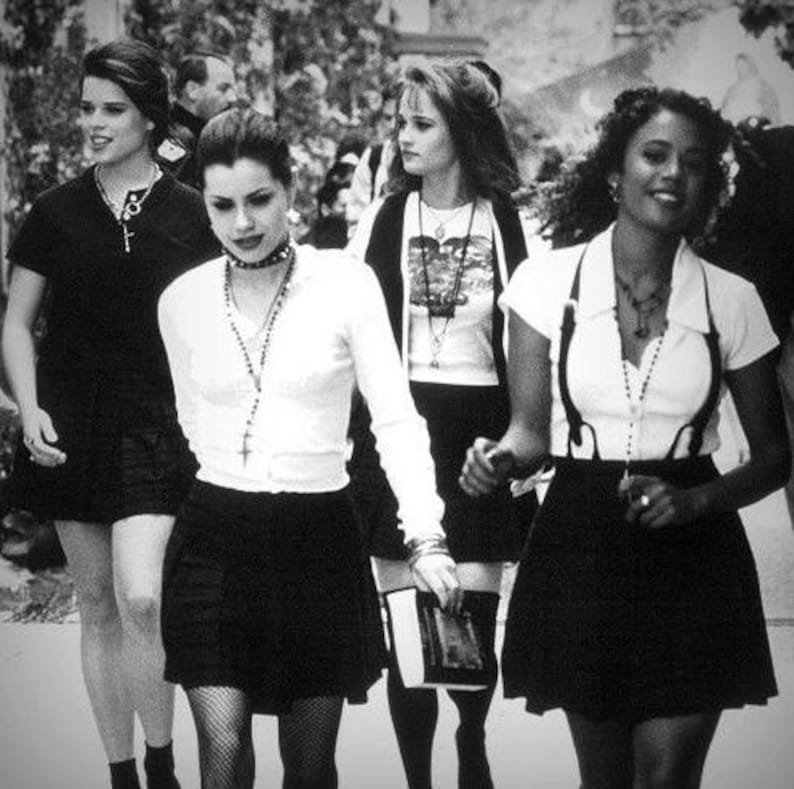 May include: Four individuals wearing black skirts and white tops, including collared shirts and cardigans. Accessories include layered necklaces, a choker, and suspenders. The scene is captured in black and white, showing a group walking together.