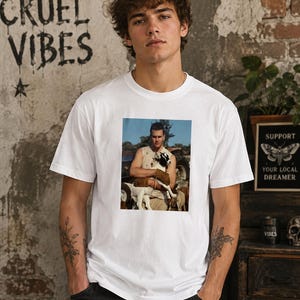 May include: A white short-sleeved t-shirt featuring a rectangular graphic print on the chest. The print depicts a person wearing a sleeveless top and gloves, holding a small goat in an outdoor setting with a vehicle visible in the background.
