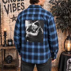 Puede incluir: Una camisa de franela de manga larga con botones en un patr&oacute;n de cuadros azul y negro. La parte posterior de la camisa presenta un gran estampado gr&aacute;fico cuadrado en blanco y negro que representa a una persona abrazando a una figura enmascarada.