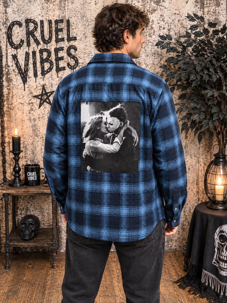 Puede incluir: Una camisa de franela de manga larga con botones en un patr&oacute;n de cuadros azul y negro. La parte posterior de la camisa presenta un gran estampado gr&aacute;fico cuadrado en blanco y negro que representa a una persona abrazando a una figura enmascarada.