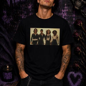 K&ouml;nnte beinhalten: Ein schwarzes T-Shirt mit kurzen &Auml;rmeln und einem rechteckigen Grafikdruck von vier Personen, die in einer Reihe stehen. Das Shirt besteht aus einfarbigem schwarzem Stoff und zeigt ein klares, zentriertes Foto der Gruppe auf der Brust.