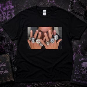 以下が含まれることがあります： 中央に長方形のグラフィックプリントが施された黒の半袖Tシャツ。プリントには顔の前にかざされた両手が描かれており、それぞれの指には石が埋め込まれた精巧な装飾が施された大きな銀色の指輪がはめられています。