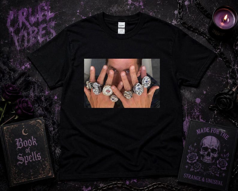 以下が含まれることがあります： 中央に長方形のグラフィックプリントが施された黒の半袖Tシャツ。プリントには顔の前にかざされた両手が描かれており、それぞれの指には石が埋め込まれた精巧な装飾が施された大きな銀色の指輪がはめられています。