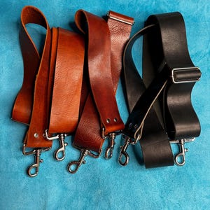 May include: Three leather bag straps in tan, reddish-brown, and black. Each strap features silver-toned metal swivel lobster clasps at both ends and an adjustable sliding buckle. The leather has a visible grain texture and is secured with metal rivets.