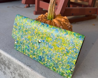 May include: A rectangular clutch wallet featuring a vibrant, speckled paint pattern in shades of bright yellow, lime green, and deep blue. The wallet has a fold-over flap secured with a single silver-toned metal snap button and silver rivets.