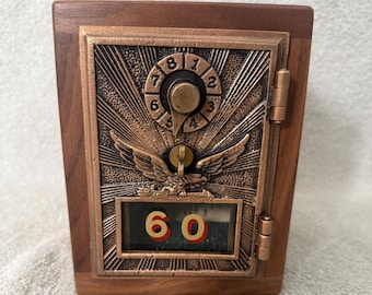 May include: A wooden box with a bronze-colored metal front resembling a vintage postal box. The metal front features an eagle emblem, a dial with numbers, and the number "60" displayed in a small window. The box is rectangular with a hinged door.