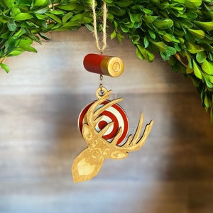 May include: A wooden deer head ornament with a red and white target in the background. The ornament is hanging from a gold string and a red and gold shotgun shell.