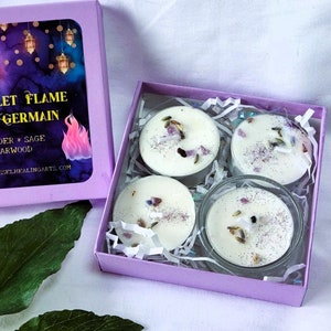 May include: A set of four lavender, sage, and cedarwood scented tea lights in a purple box. The box has a label that reads "Violet Flame St. Germain Lavender * Sage Cedarwood Sacredvesselhealingarts.com".